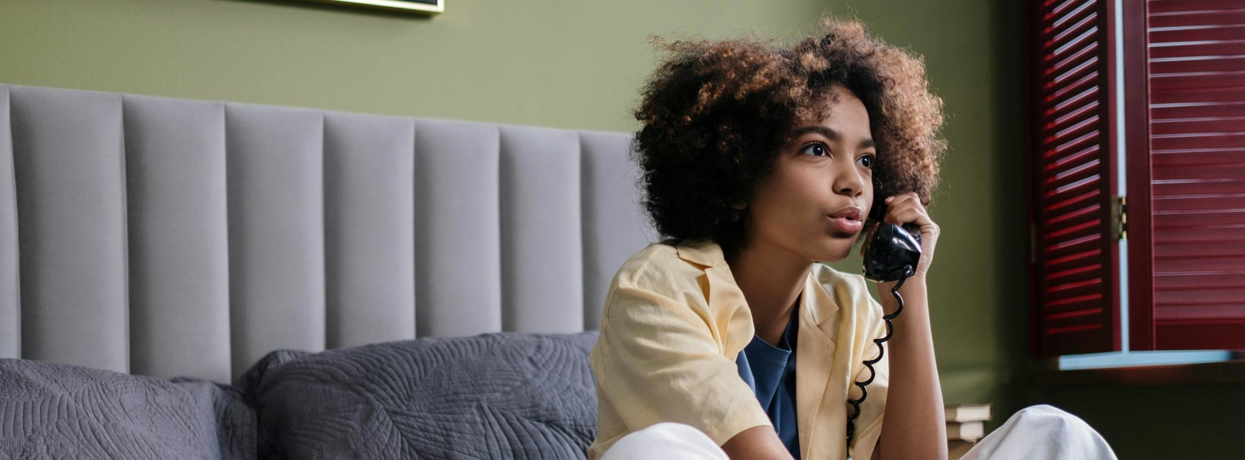 Image shows a young woman sitting on a bed and talking on a landline telephone.